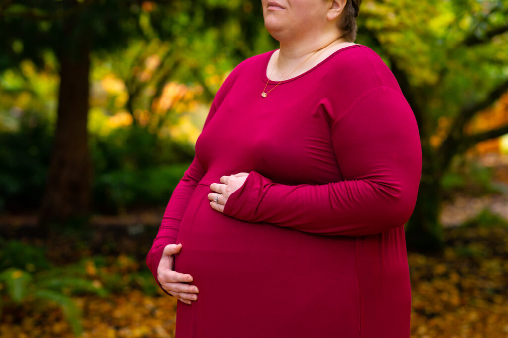 A plus-size woman with pale skin, short brown hair and glasses stands in the middle of fallen yellow leaves with autumn trees and bushes around her. She is wearing a necklace, long pink dress, sandals and flower crown, and is cradling her belly with her hands. She's looking away from the camera with a peaceful or pensive expression; only her lower face, arms, torso and belly are shown.